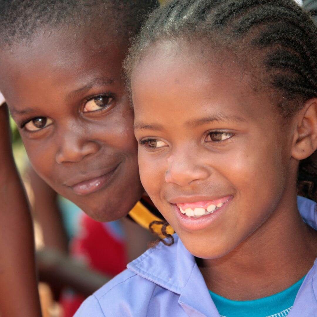 Zwei Kinder mit dunkler Hautfarbe lächeln fröhlich in die Kamera. Das Mädchen im Vordergrund trägt eine hellblaue Jacke und hat geflochtene Haare. Der Junge hinter ihr, und lehnt sich leicht zu ihr hin. Beide lächeln.
